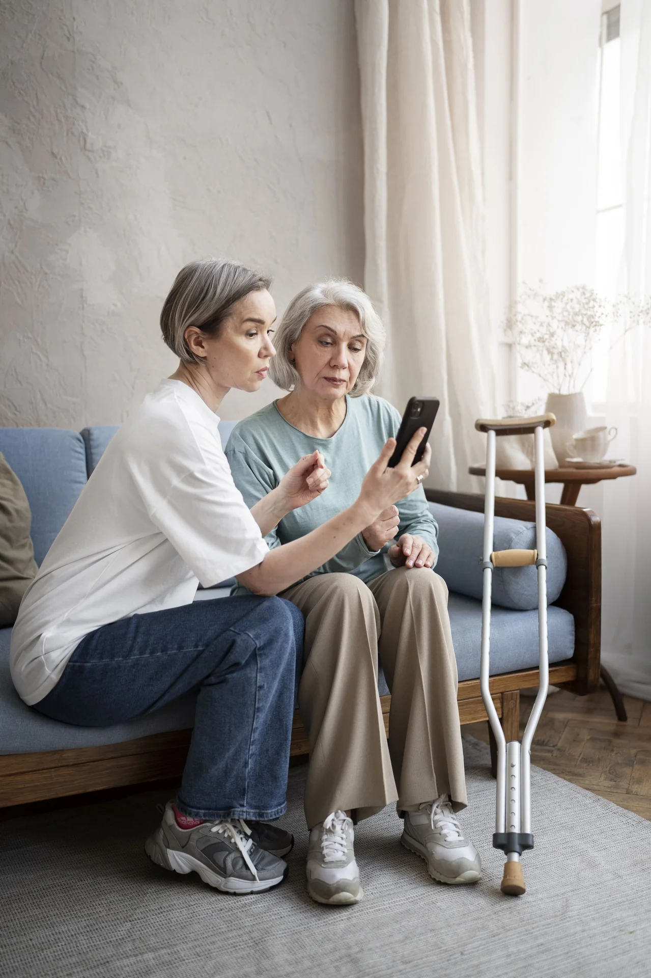 Dos mujeres sentadas en un sofá consultando un teléfono móvil, con una muleta apoyada a su lado.