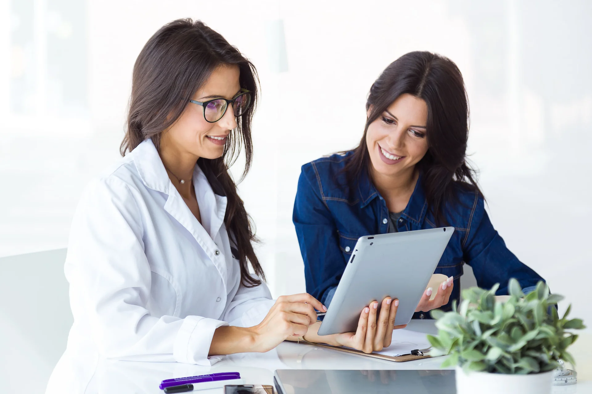 Doctora con bata blanca y una mujer consultando juntas una tableta.